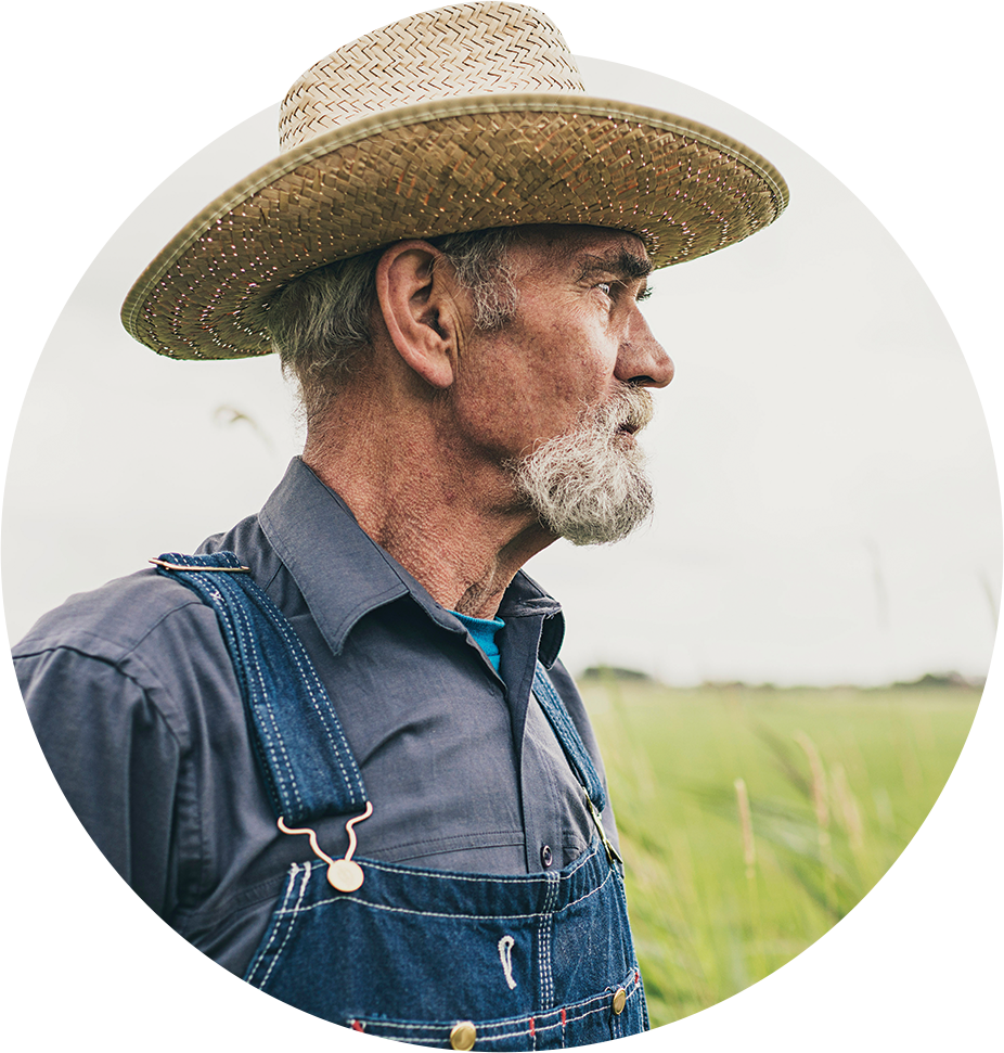 Side profile portrait of an older male farm worker wearing a straw hat, grey shirt, and blue overalls in front of a field looking into the distance to the right.